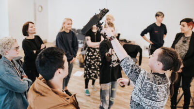 A white person with long dark hair and a funky jumper holds up a boom mic and points at it, whilst other stand round in a circle, an attentive audience.