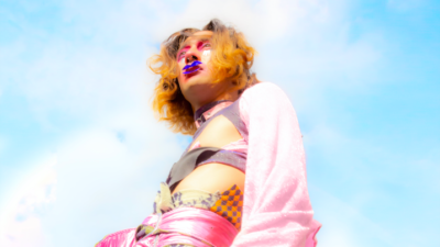 A tall, thin person photographed from below. They wear yellow and purple patterned trousers cinched by a shimmery wise pink belt, a purple bralet too and flowing pink sleeves that extend past their hands. They have short curly hair which is blonde and brown, and wear pink eyeshadow and a painted blue moustache. They look thoughtfully above the camera and the backdrop is a cloudy, dreamy blue sky.