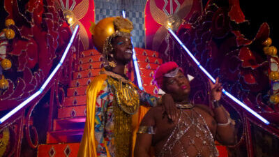 Two Black people with queer, Afrofuturist gold, silver, and multi-coloured outfits stand at the bottom of a set of futuristic, theatrical red stairs. One person leans on the other's shoulder, who holds up their fingers in a peace sign.