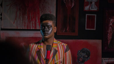 A Black person with a black circle painted on their face and wearing a colourful stripy jacket is in a room with red paint splattered on the walls and a microphone in front of them.