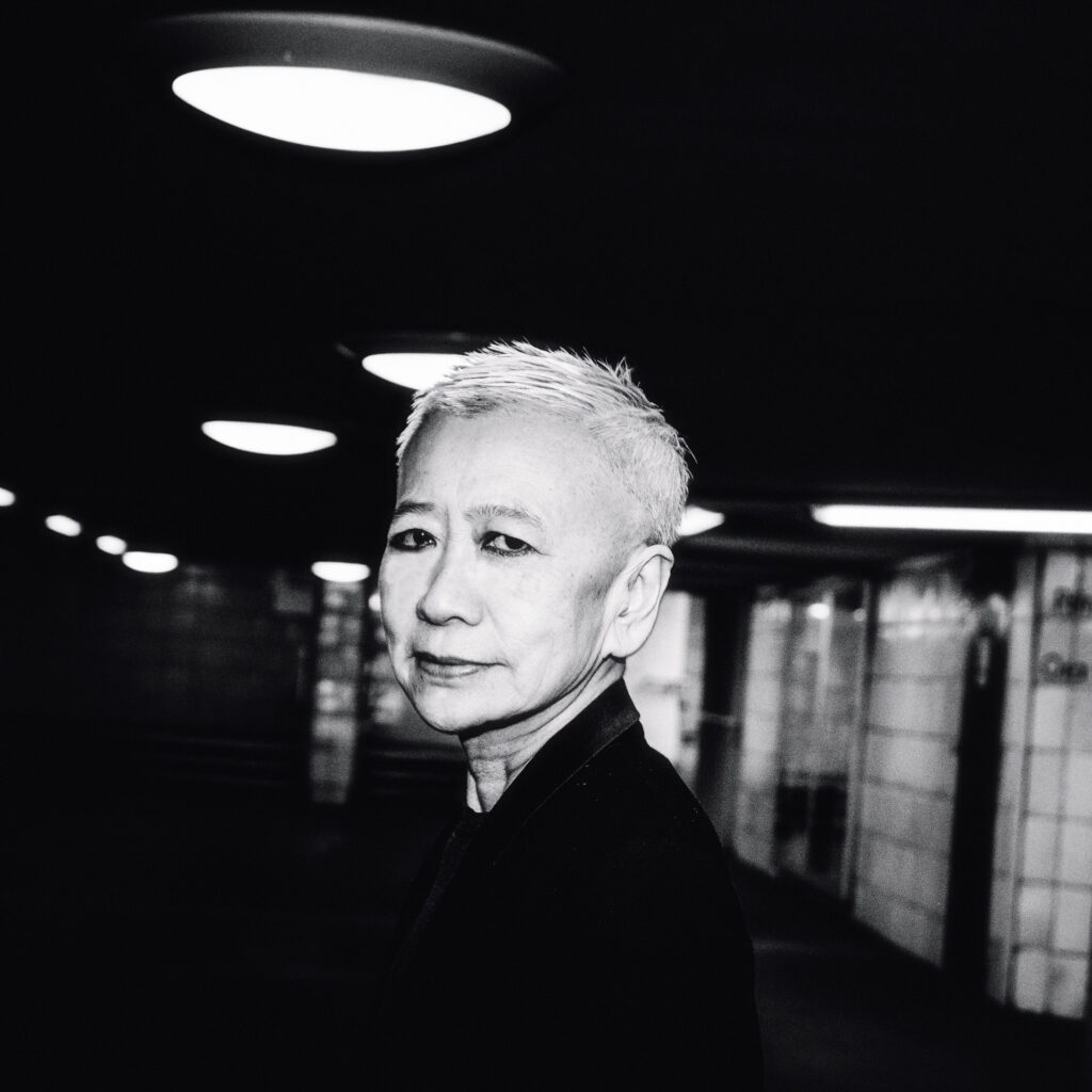 A black and white headshot of Shu Lea Cheang, a Taiwanese-American woman with short bleach blond hair, standing in a darkened corridor with bright overhead lights.