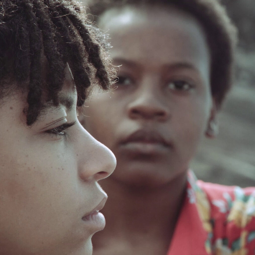 Close-up side profile of a young Black person looking ahead of them, whilst behind them another young Black person with a colourful shirt looks at the first person.