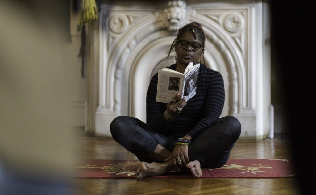 An African American woman sits cross-legged on a yoga mat, reading from a text, which you can make our an image of poet Audrey Lorde on the cover over. The woman is wearing a stripy black top and shiny black leggings, and colourful bracelets and rings. She wears dark glasses and has braided hair. In the background, there is an ornate white fireplace. The foreground has some blurry objects which suggest others sitting and listening to her read.