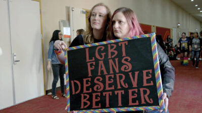 Still from the film Queering the Script in which two white people standing in a hall at a fan convention hold up a sign which reads, "LGBT fans deserve better."
