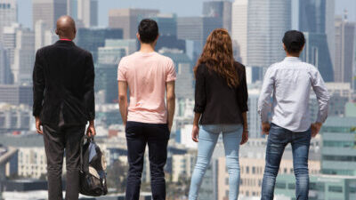 Four people stand with their backs to us, facing the city of San Francisco with tall buildings and blue sky in front of them.