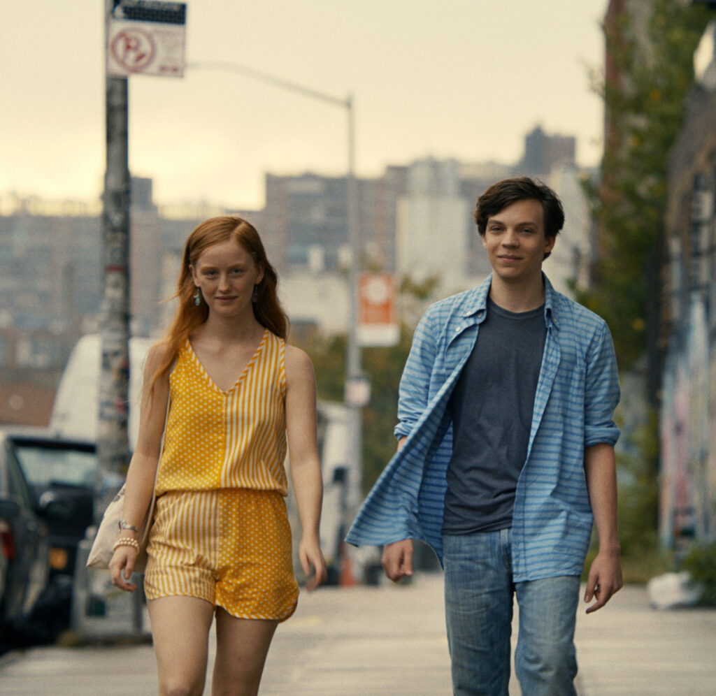 Two young people walking down a city street. One is a white woman wearing a yellow jumpsuit, and the other is a white man wearing a blue shirt and jeans. walking down a city street. They look purposeful and excited. 