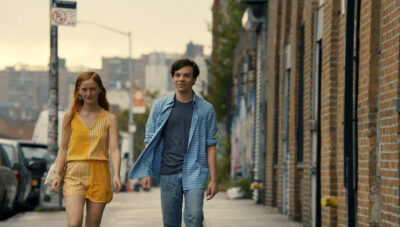 Two young people walking down a city street. One is a white woman wearing a yellow jumpsuit, and the other is a white man wearing a blue shirt and jeans. walking down a city street. They look purposeful and excited.