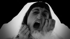 A close up black and white image of a person's face. They are clawing at a clear veil covering their face and have their mouth open like they are yelling. They look angry, desperate and fierce.