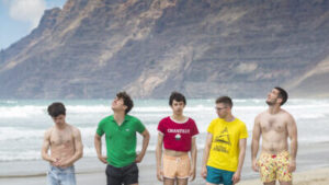 Five people, all with short hair and wearing different kinds of shorts, some swimming togs, one person in jean shorts, stand in a line-up on a beach, with the sea coming in behind them. Behind the waves, there are volcanic mountains covered with clouds. One person in the centre of the group, wearing a red t-shirt with the word 'CHANTILLY', rolled up to reveal their stomach, is staring into the camera. The others, in colourful tops or topless, are looking at the sky, or downwards.