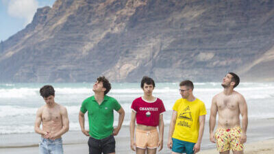 Five people, all with short hair and wearing different kinds of shorts, some swimming togs, one person in jean shorts, stand in a line-up on a beach, with the sea coming in behind them. Behind the waves, there are volcanic mountains covered with clouds. One person in the centre of the group, wearing a red t-shirt with the word 'CHANTILLY', rolled up to reveal their stomach, is staring into the camera. The others, in colourful tops or topless, are looking at the sky, or downwards.