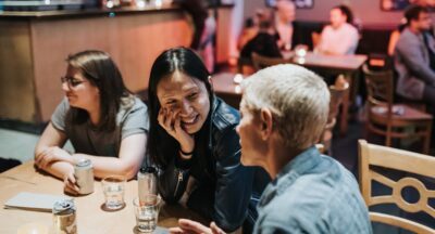 three people sitting at a table in the CCA bar, with mellow lighting. A woman with long black hair sits smiling listening to a non binary person with short blonde hair talking. The third person is looking to the rest of the table. In the background others are talking at other tables. The mood is friendly and social.