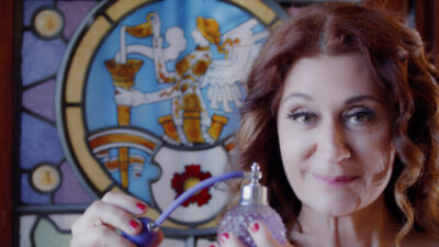 A person with long, wavy auburn hair with highlights, with long eyelashes and pink lipstick, smiles at the camera, and is holding a beautiful vintage perfume bottle with a purple tassel. Behind them is a painted stained glass window, with a picture of a figure with wings and a hat holding a gold horn-like object with a ghost-like figure emerging from the object.