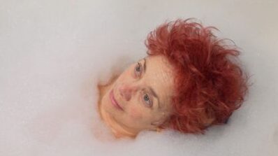 A white nonbinary person with red hair lying in the bath looking up at the ceiling. They are surrounded by bubbles. They look zoned out.