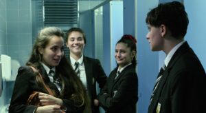 Three school girls stare at a boy with vicious smiles on their faces. One of the girls is tightly holding a brown school bag. They are in a school bathroom, and the boy looks worried and upset.