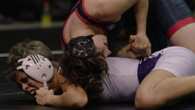 A close up of two white people wrestling. One is laying on top of the other, who is shouting. They both wear protective head gear and sports tunics. The image feels dynamic and active.