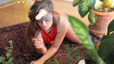 A person with long brown hair crawls on a persian-style rug in a room with wood-laminate floors, surrounded by green leafy potted plants. They are blindfolded, and have a dildo strapped to their forehead.