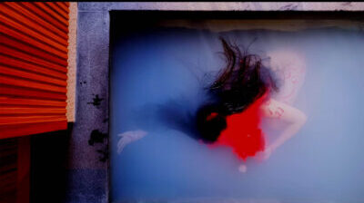 A person is submerged in a pool of milky water . The person looks in a state of undress, and has long black hair. Their face is covered by a thick, bright red liquid, which appears to be coming from their head and gathering around their body. To the left of the image is a red wood panelled wall.