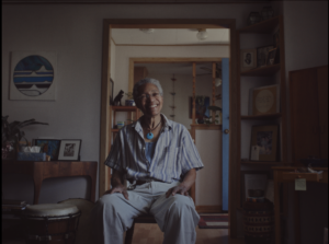 an older black man with white hair sits in his house, with a big smile. He is wearing a blue stripy shirt which is unbuttoned halfway, a blue stone necklace and cream trousers. The house looks well-lived in and the image is warm.