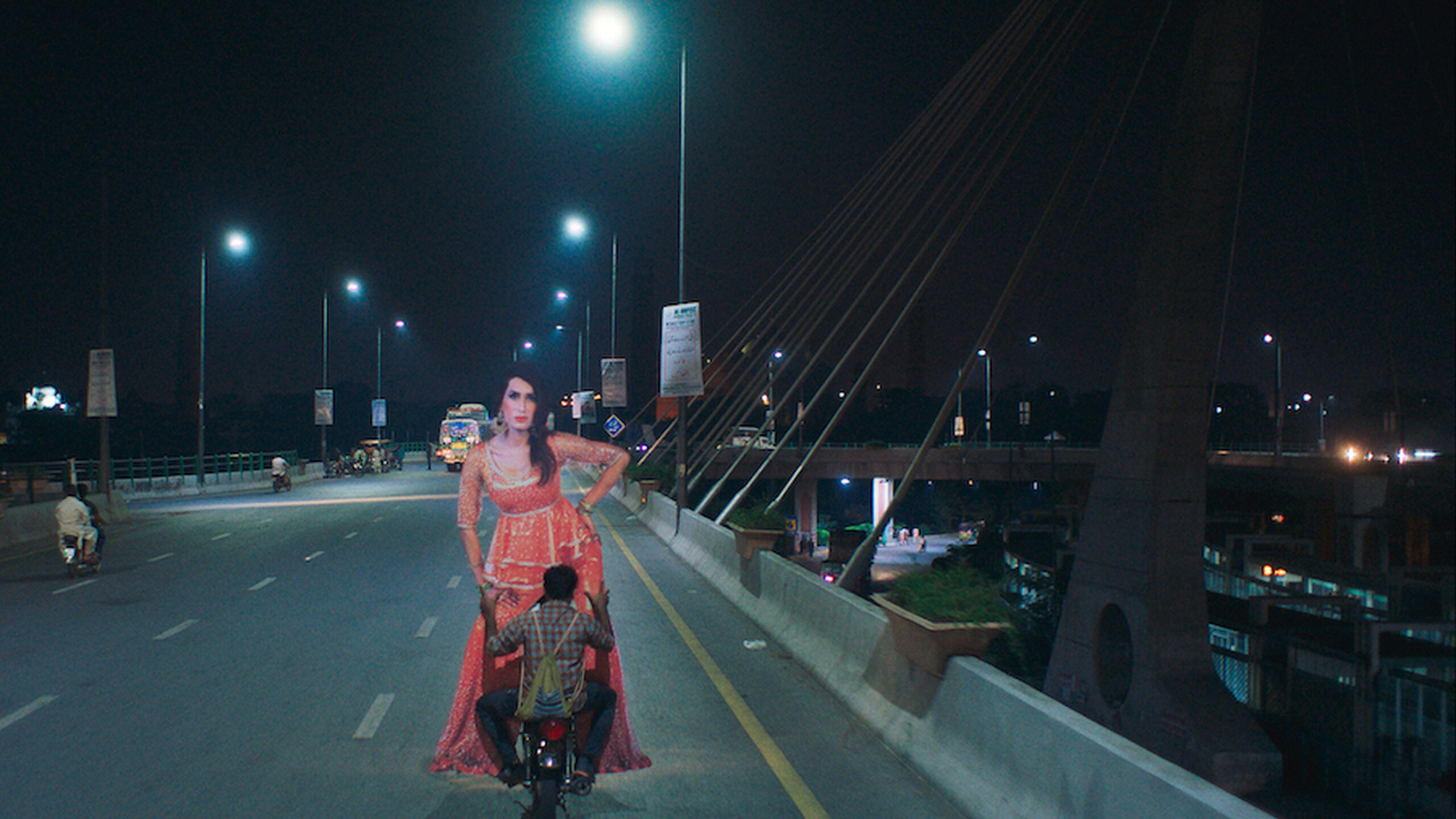 A night scene of a man holding a large cut out of a woman whilst riding a moped across a bridge.