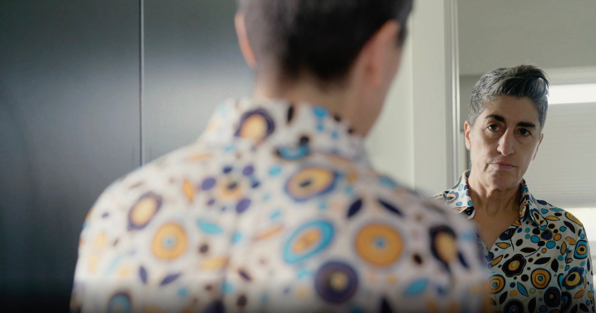 Sarah looks at herself in the mirror. She’s wearing a colourful shirt with designs in blue, orange and purple.