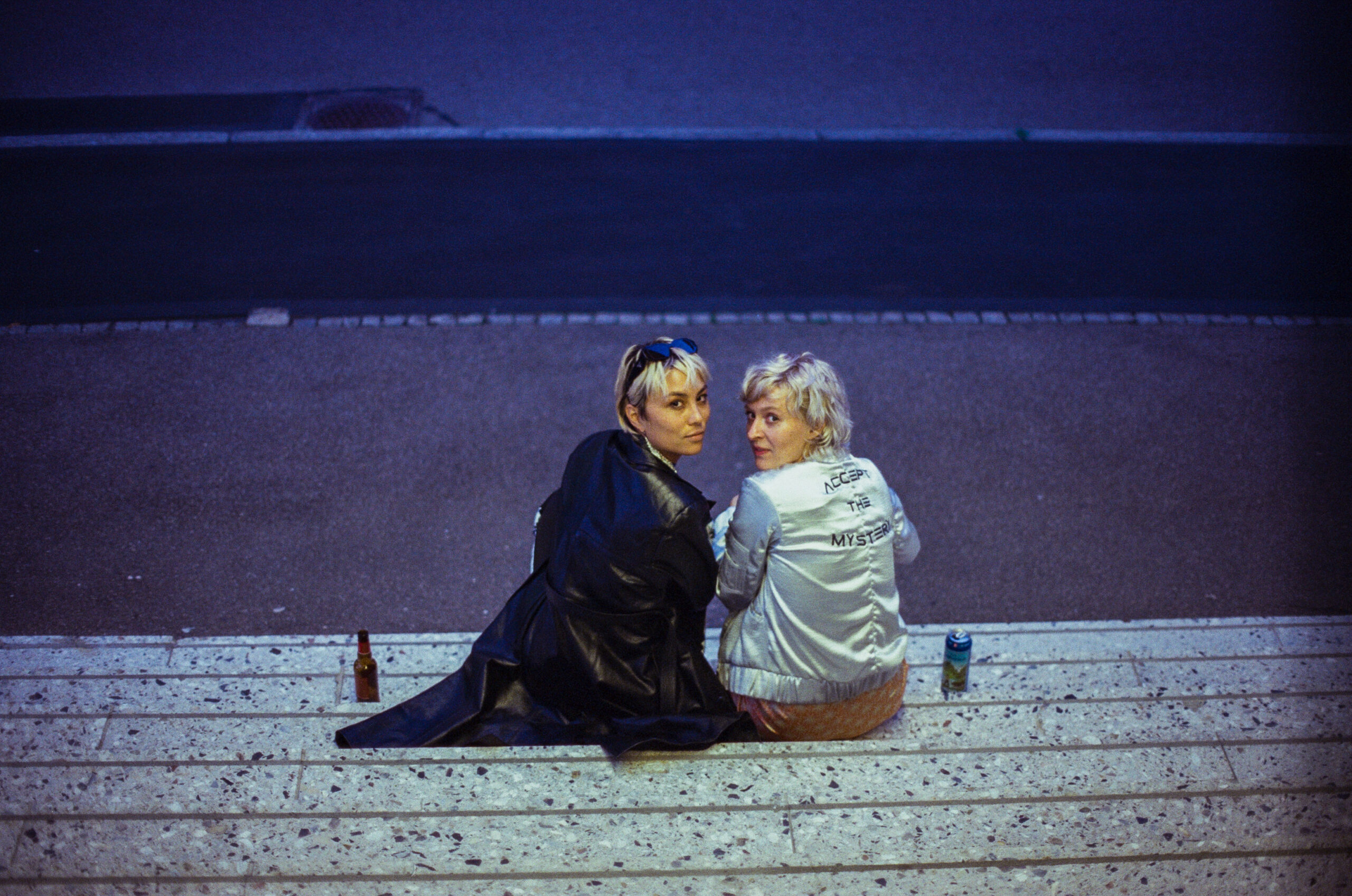 A street image with blue lighting featuring two women in the middle of the shot seating down at some stairs and looking up to the camera.