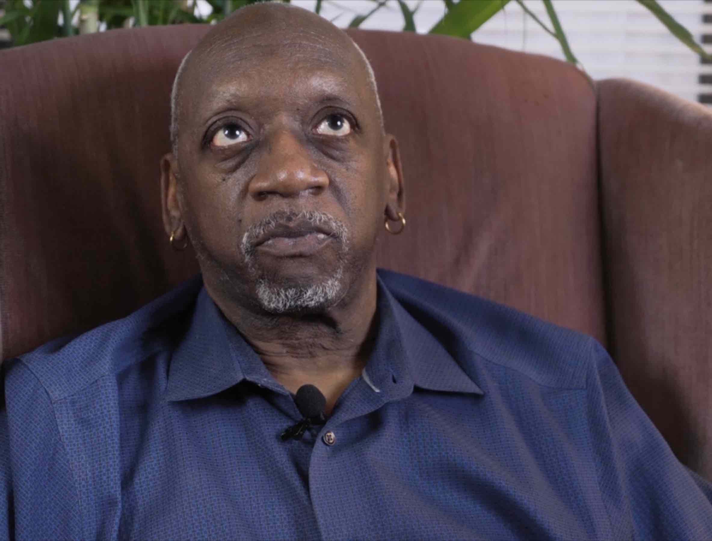 Cornelius, a Black man wearing a blue shirt and sitting on a brown sofa looks at the camera.