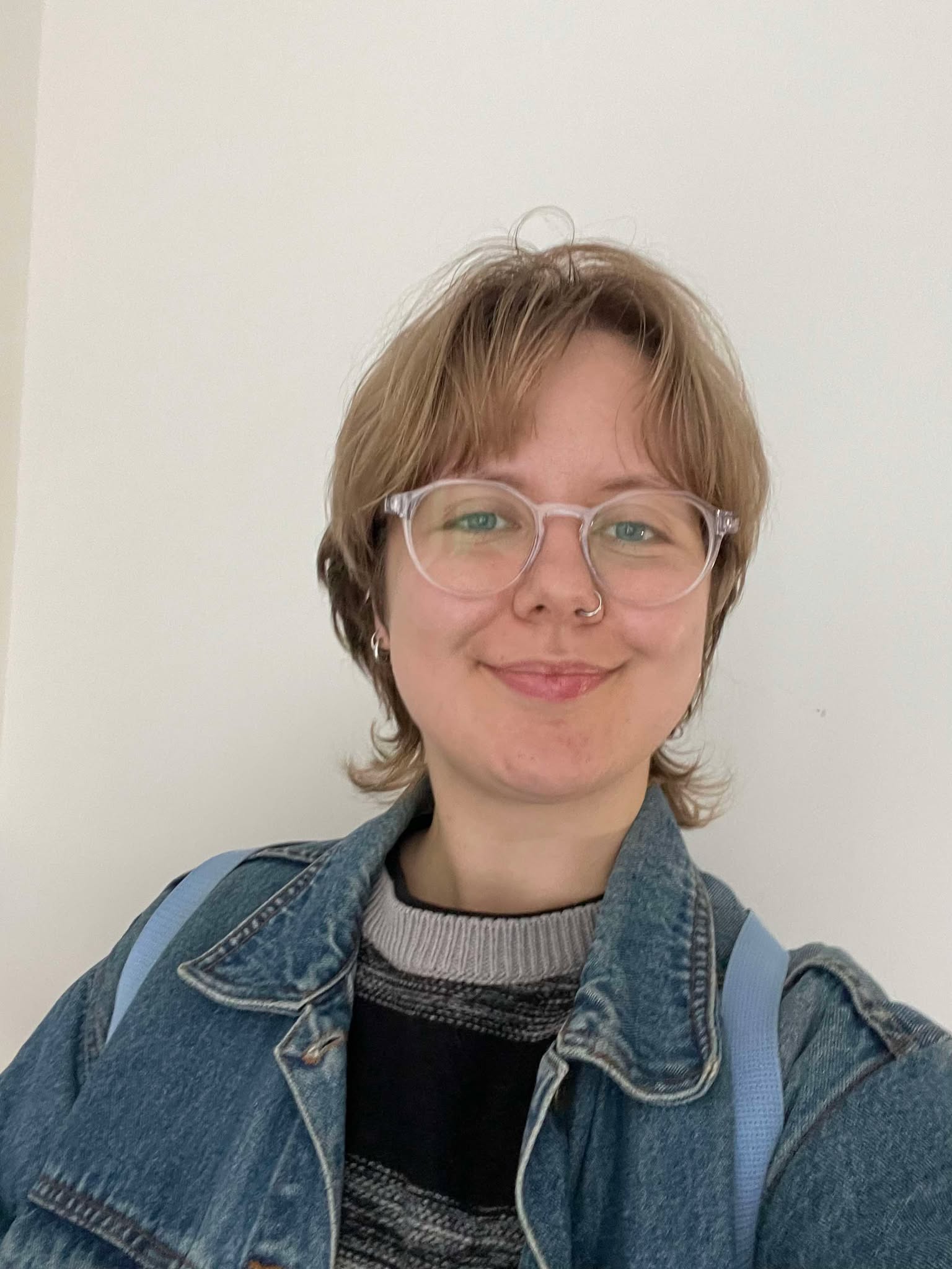 A selfie of a smiling white person with short brown hair, a nose piercing and clear-framed glasses. They are wearing a striped jumper and a denim jacket. The background is white.