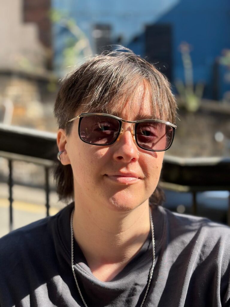 Erica, a white non-binary person with dark hair and wearing sunglasses, looks at the camera