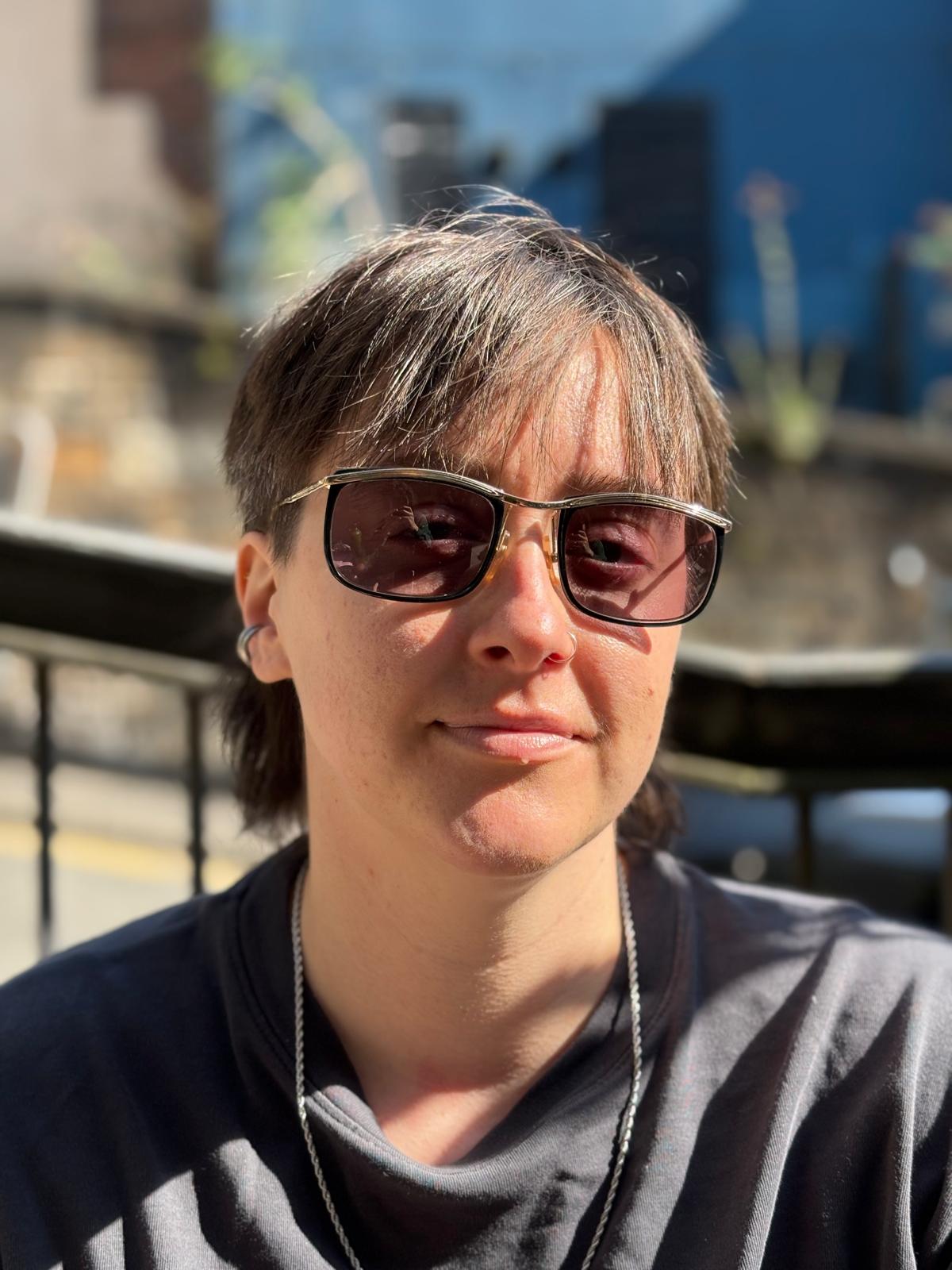 Erica, a white non-binary person with dark hair and wearing sunglasses, looks at the camera