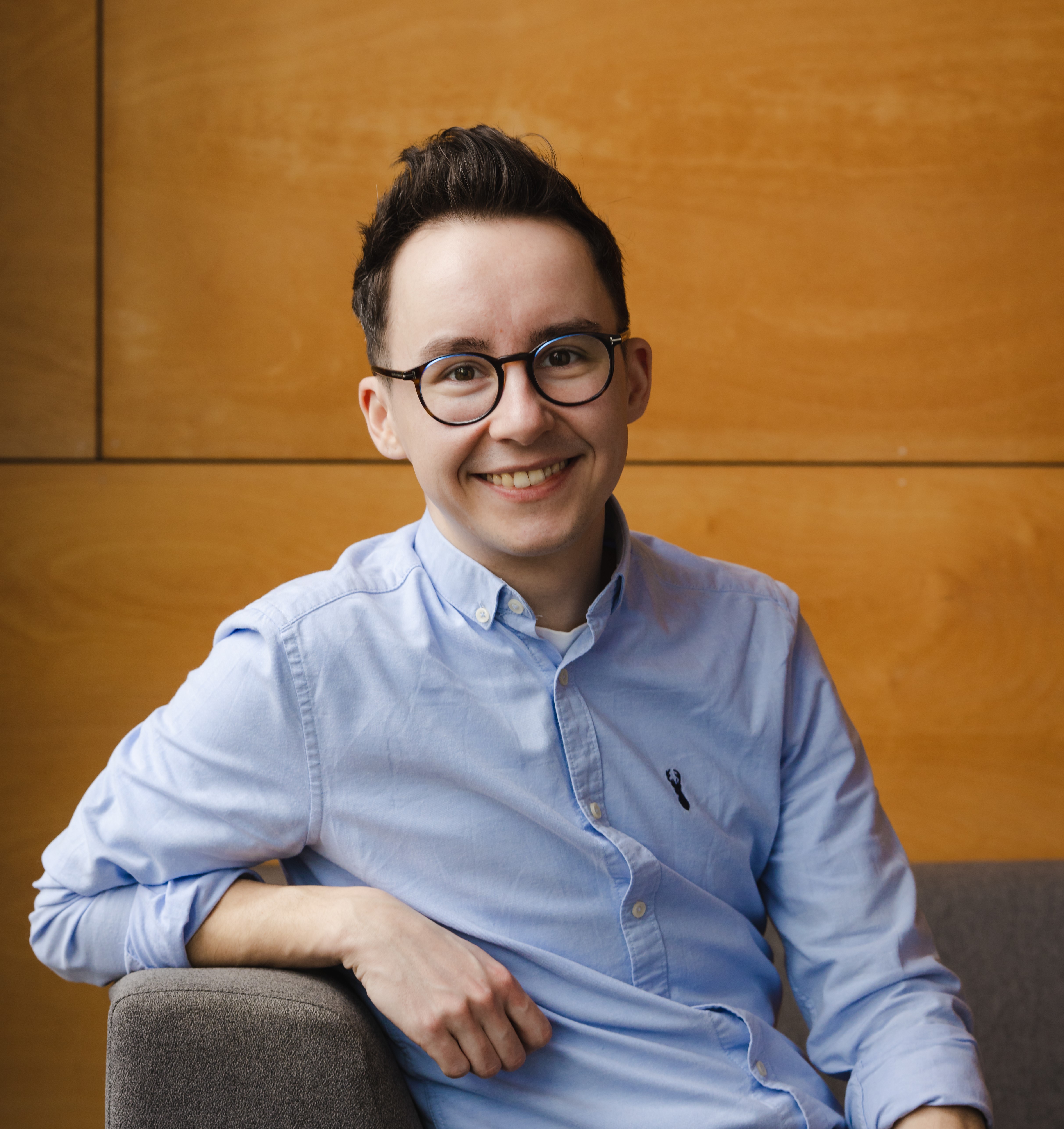 A smiling person sitting on a grey chair in front of a wood paneled background wearing a light blue shirt, dark round glasses and short dark hair.
