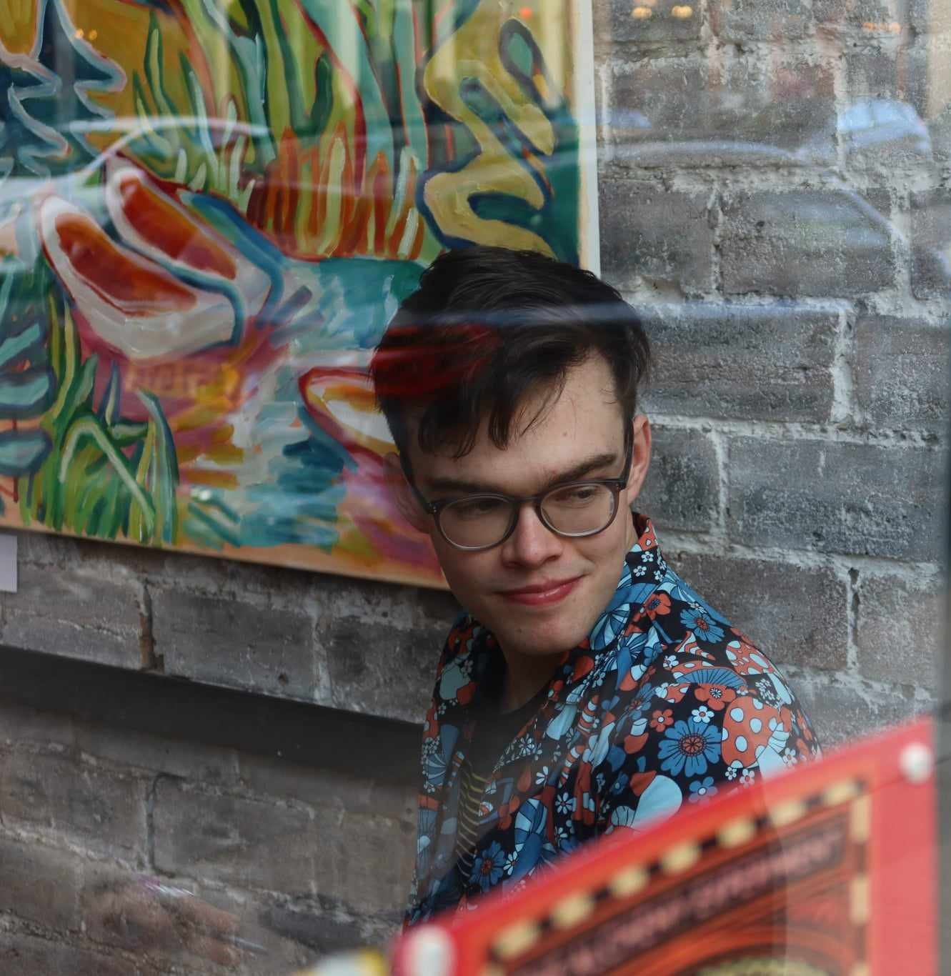 A white non-binary person with short hair and glasses sits in front of a painting and a brick wall.
