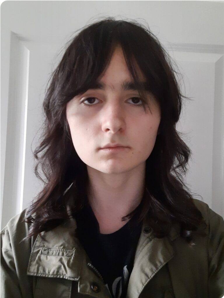 A picture of Kieran Loughran, a male presenting person with long dark hair and brown eyes, a black t-shirt and a green jacket.