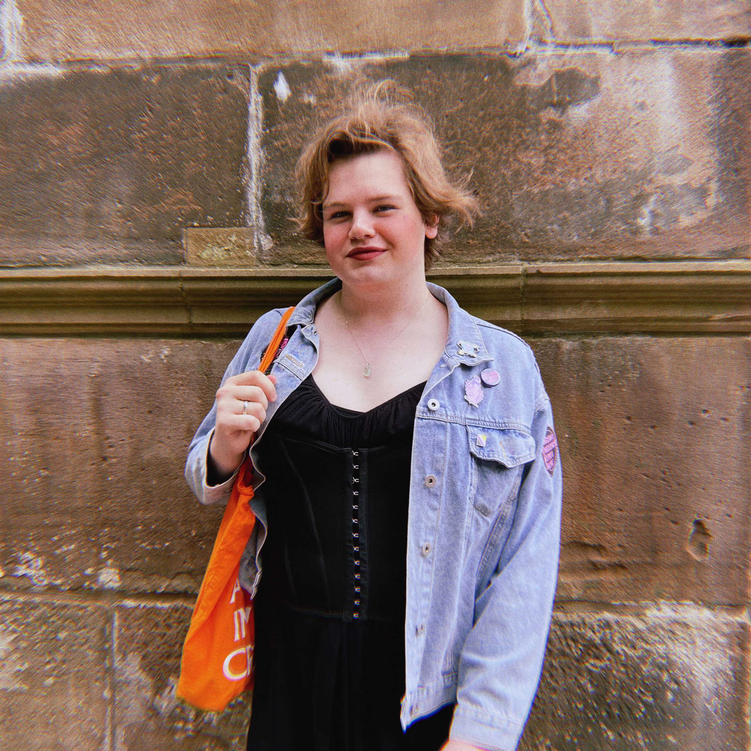 Paulie, wearing a denim jacket with badges, with neck length hair, is standing in front of a brick wall.