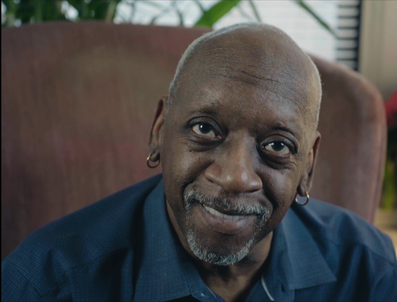 Cornelius, a Black man wearing a blue shirt and sitting on a brown sofa looks at the camera smiling.
