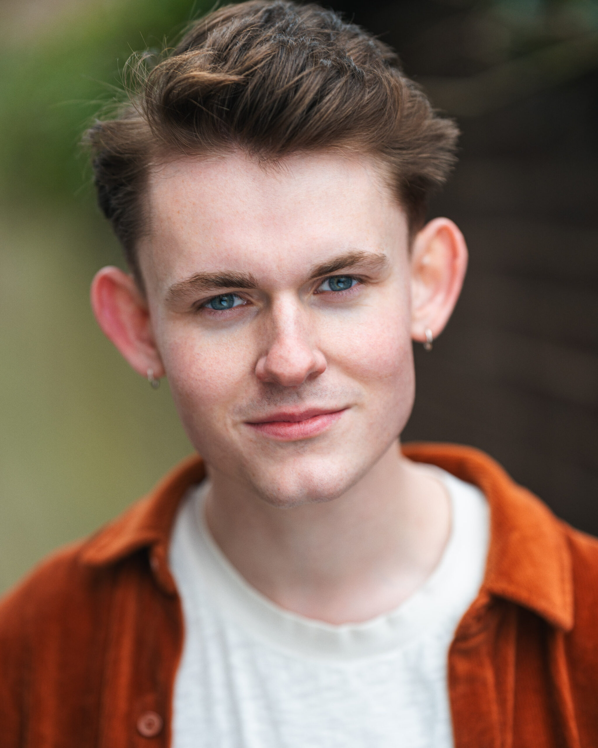 A young masculine-presenting person partially smiling at the camera while the green background is out of focus
