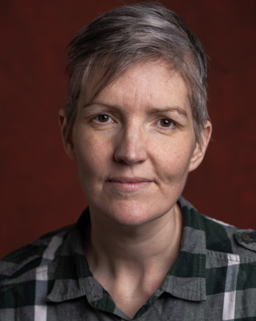 A closeup headshot of a non-binary white person in their early 40s, with short grey hair in various hues. They are looking straight ahead to the camera with a mainly neutral expression. They are wearing a green and white plaid shirt and posing against a red background.