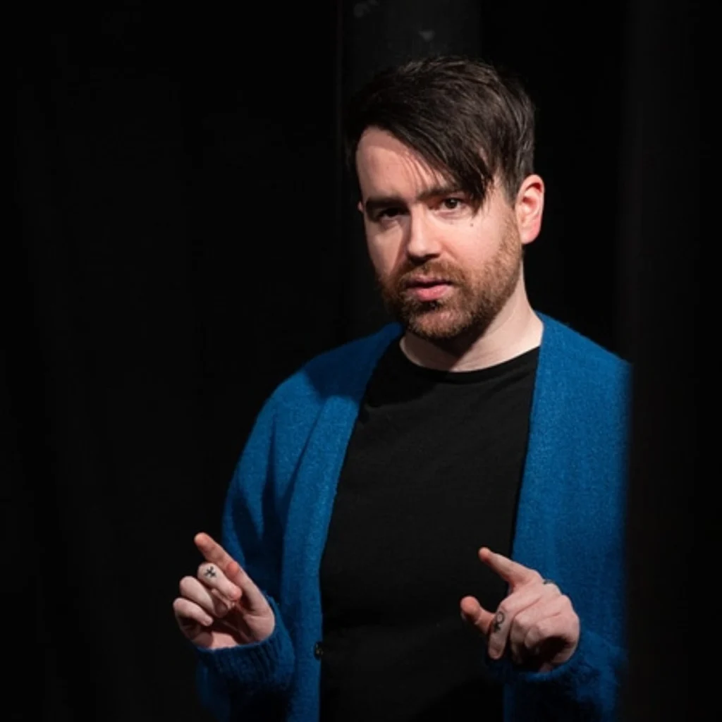 Man with hand tattoos on stage in a blue jumper, black background,