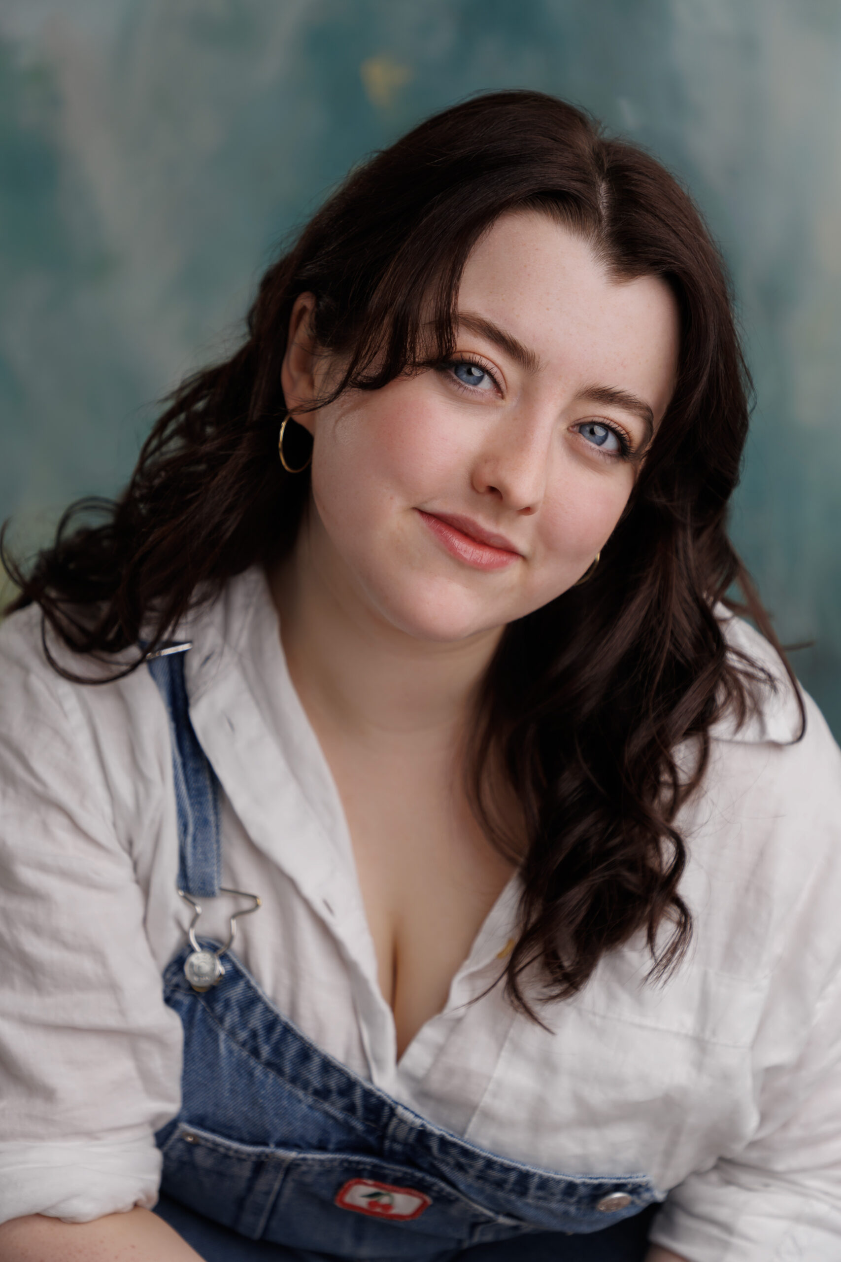 a photo of Mackenzie with dark brown hair, white skin and blue eyes. She is wearing a white shirt and blue denim dungarees. She is sat in front of a blue marbled backdrop.