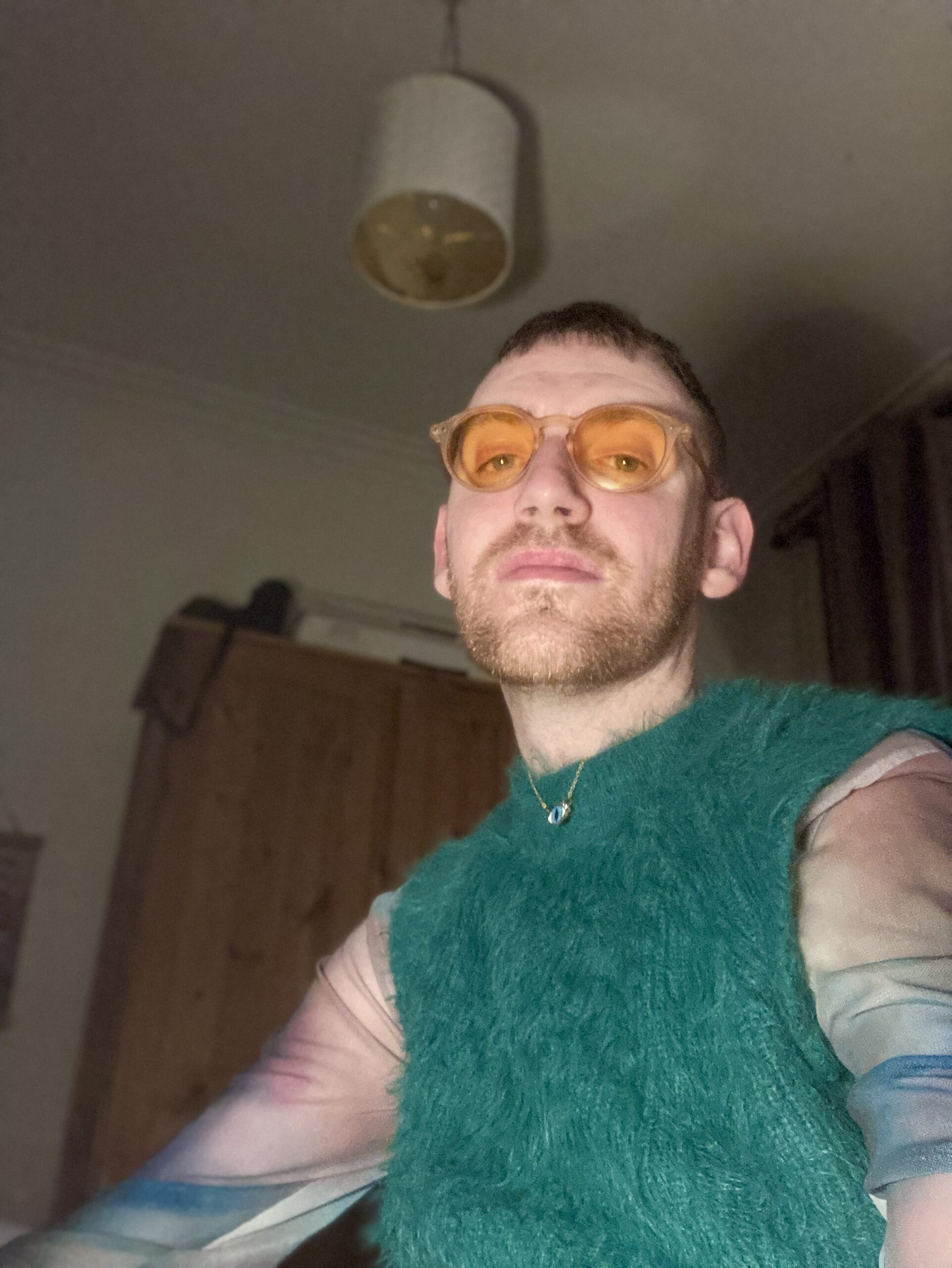 Graham Webster wears some yellow framed glasses and wears a furry green tank top. He sits in a dark bedroom.