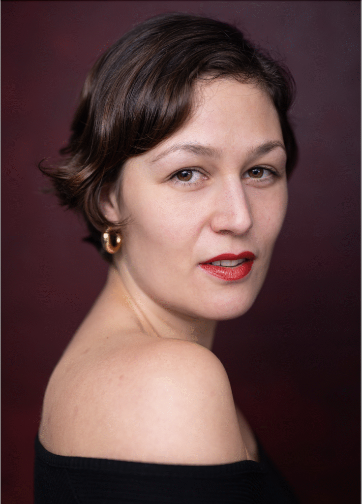 A woman with short, dark brown, wavy hair looking back over her shoulder. She has olive skin, wears red lipstick, and is smiling slightly. She's wearing an off shoulder black top.