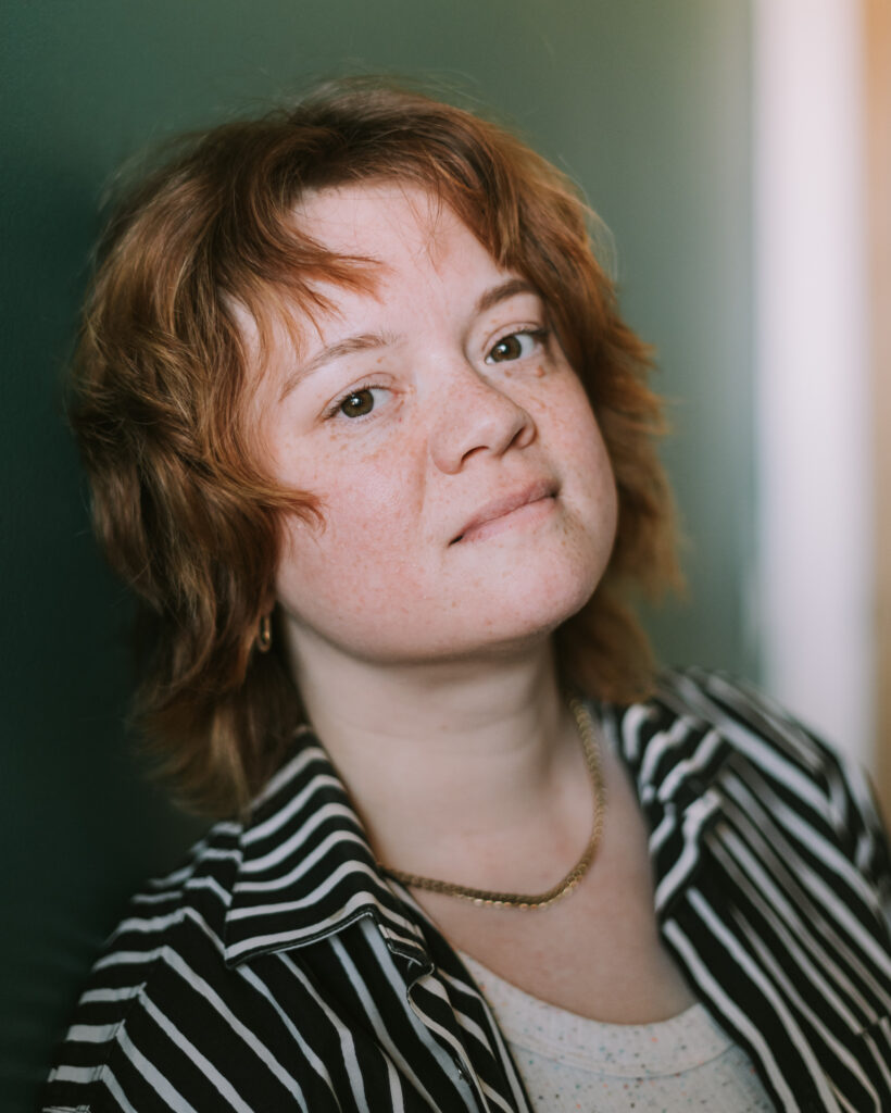 A headshot of a white person with shoulder-length, wavy reddish hair and freckles. They are wearing a striped collared shirt, white tank top and gold chain necklace. They are leaning back against a green wall and looking at the camera.