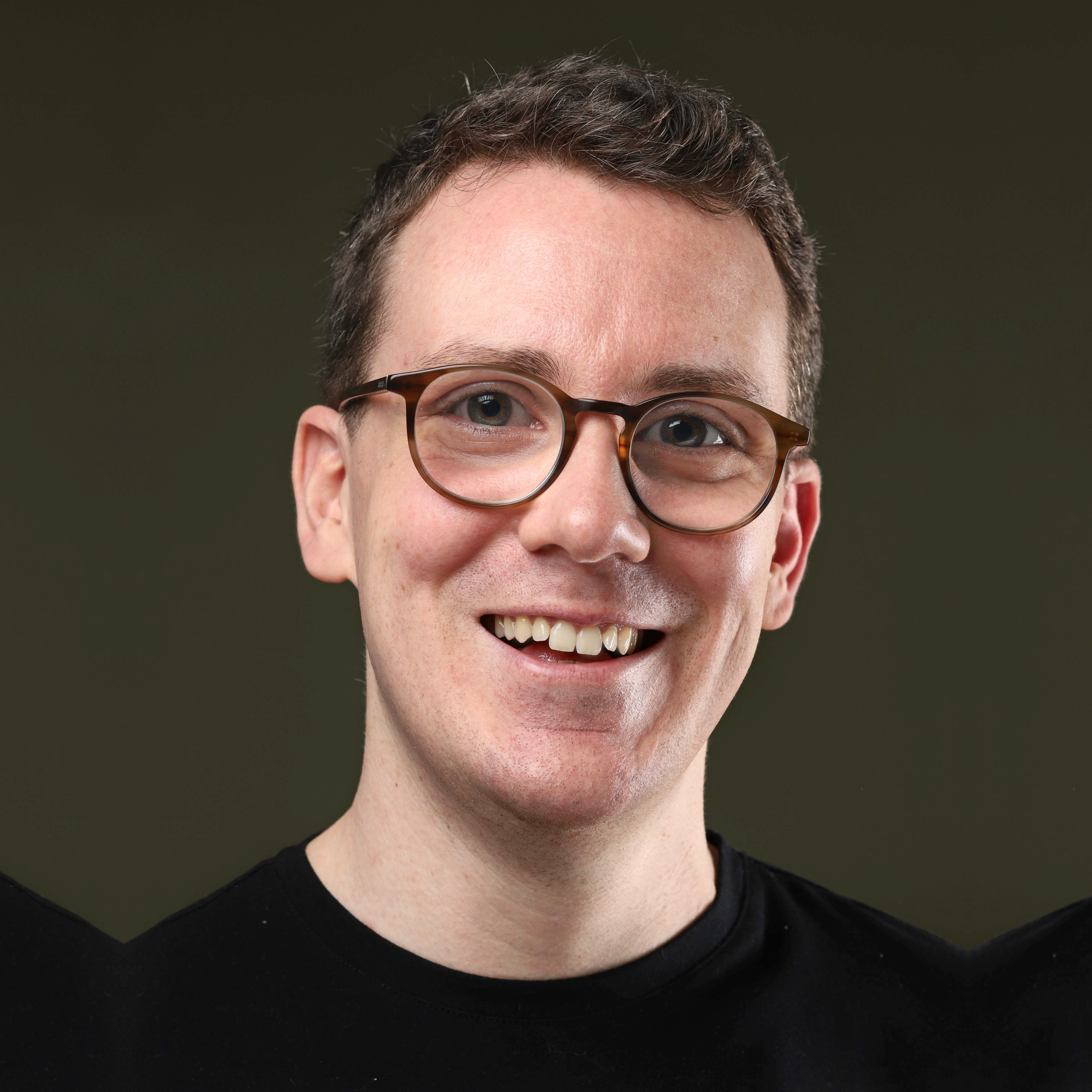A portrait of a man smiling. He has circular glasses and his eyes are magnified. He has short brown hair and wearing a black t-shirt.