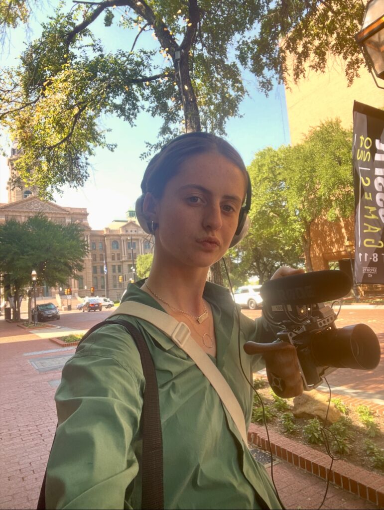 Therese poses with their camera held up in a selfie. They stand on the street in Texas wearing a green shirt with offices behind them.