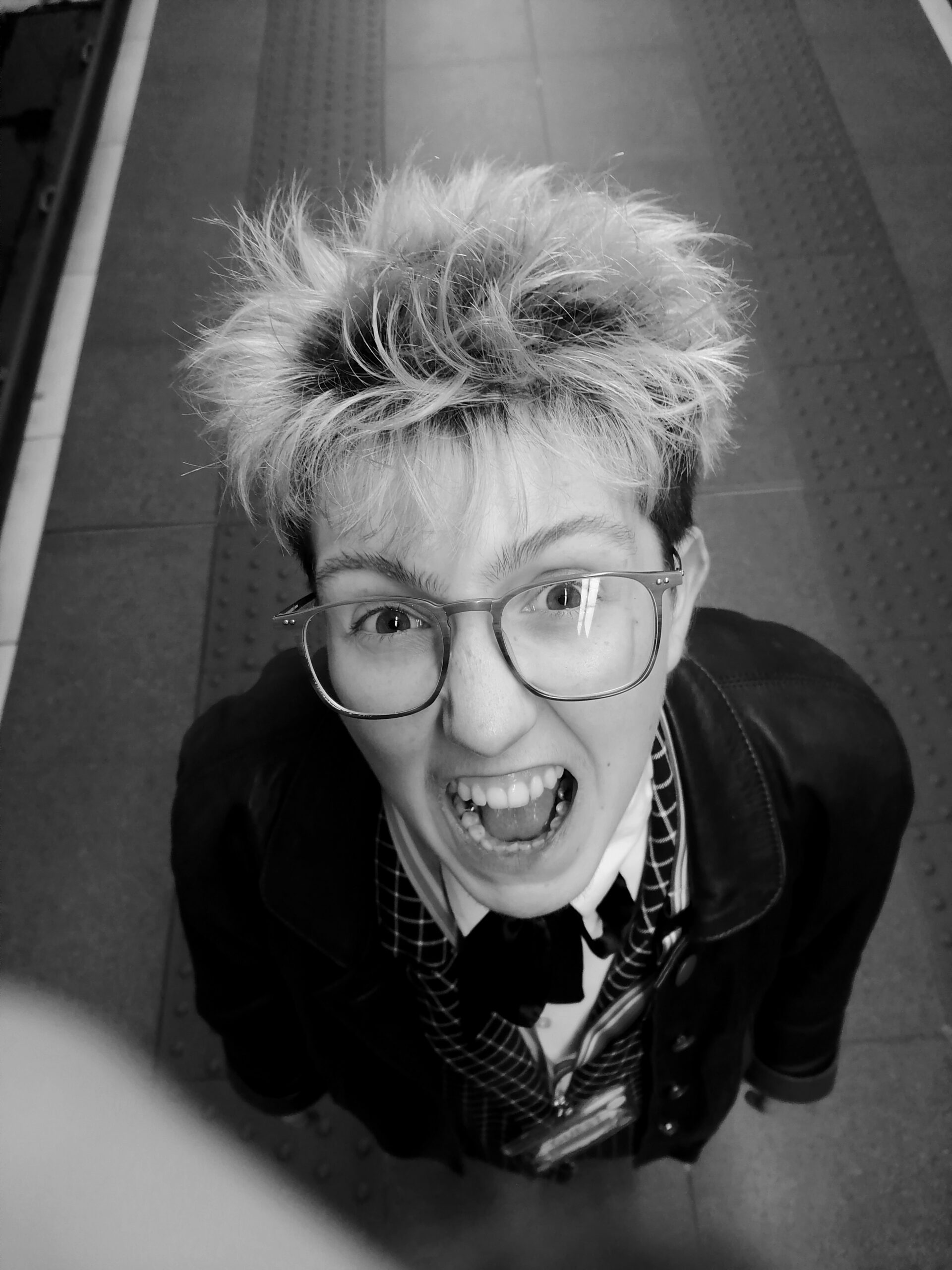 A young white person in a checkered suit with energetic hair being viewed from above, pulling a silly facial expression.