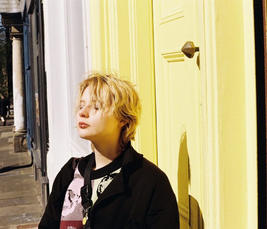 A person with short blonde hair wearing a black coat and a colorful jumper is standing with their eyes closed in front of a sun-lit bright yellow door.