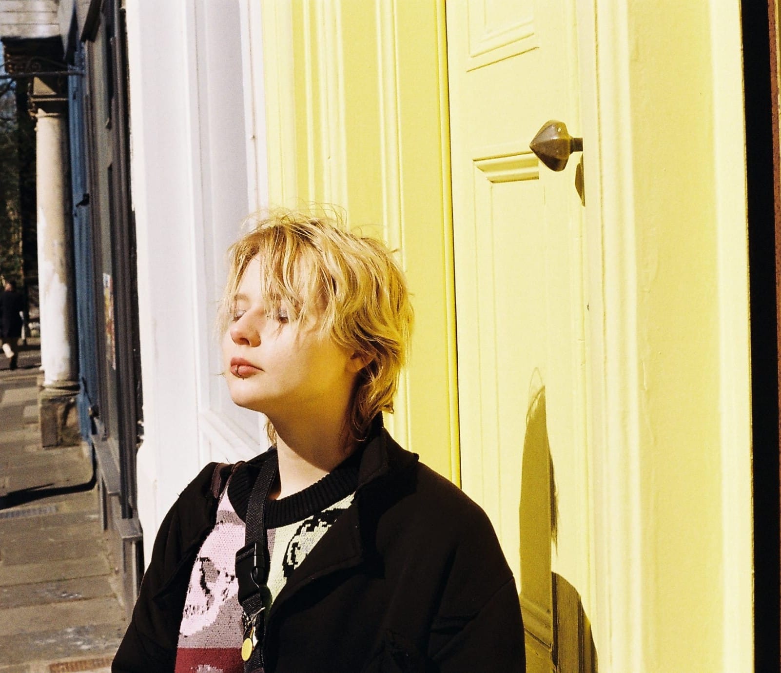 A person with short blonde hair wearing a black coat and a colorful jumper is standing with their eyes closed in front of a sun-lit bright yellow door.