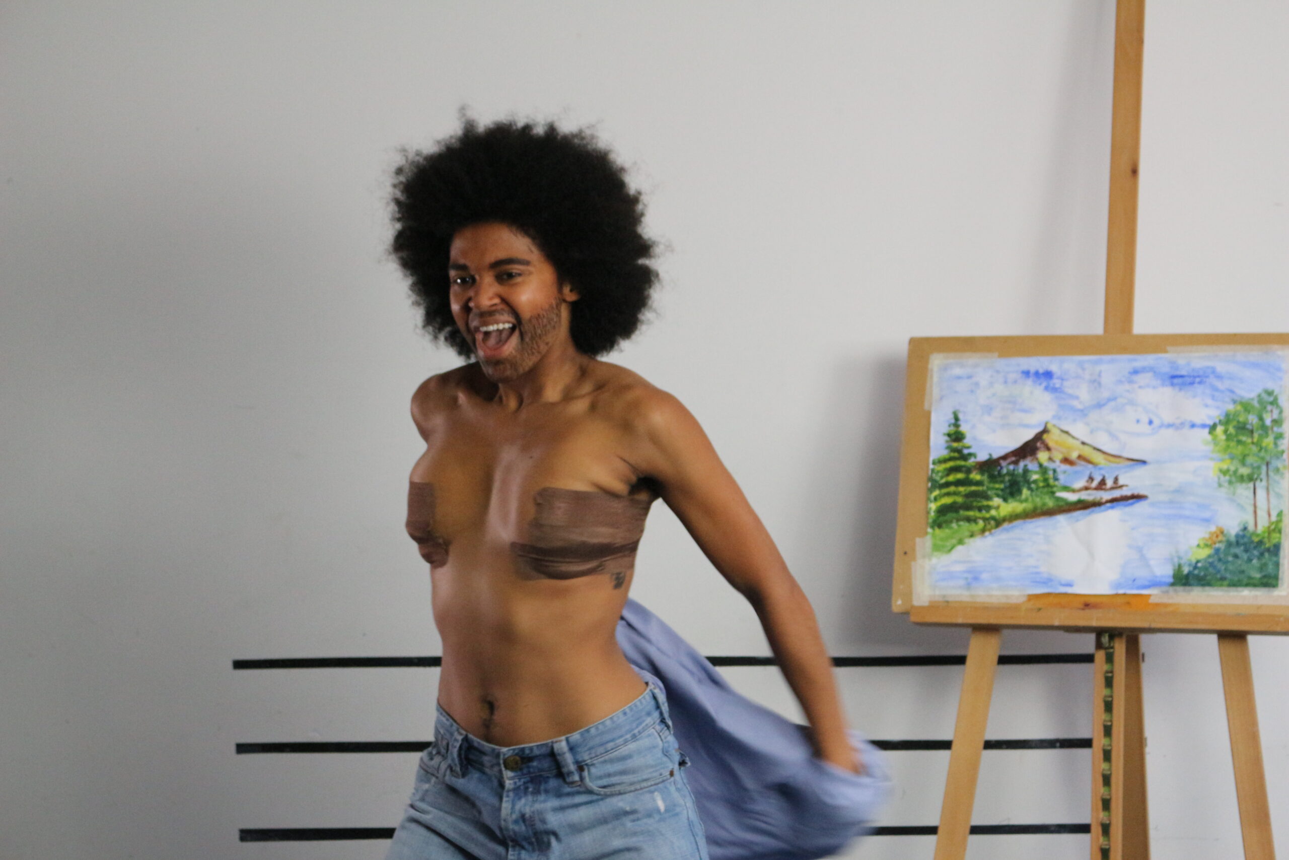 A Black drag king stands topless next to a canvas painting.