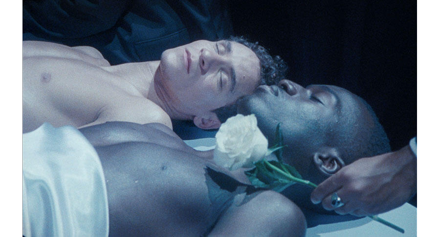 A white person and a Black person lie next to each other topless and seemingly motionless. A flower is being laid amongst them in the bottom right corner.