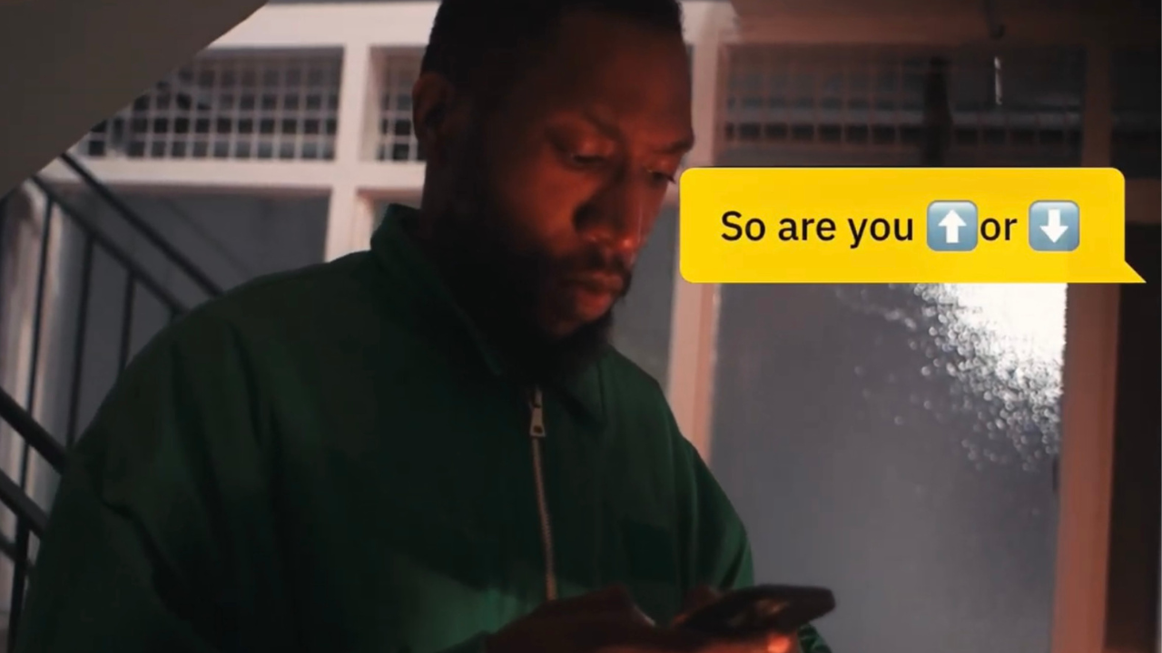 A Black man is looking at his phone and a Grindr message in on the right hand side of the screen with the text: "So are you a top or a bottom?"