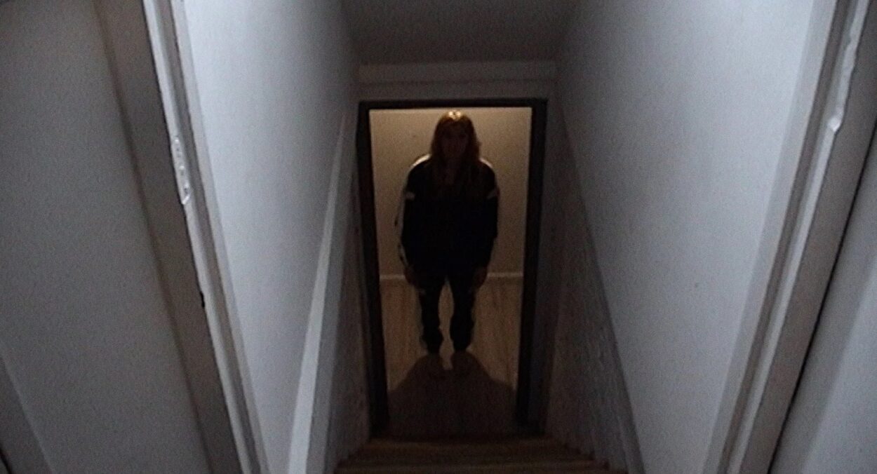 Circle, a trans white woman with long blond hair is standing in between a door and looking up the stairs.